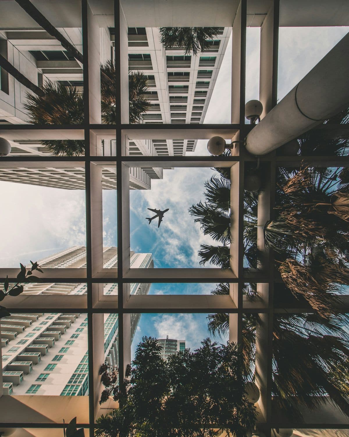 Upward view through a white grid of concrete beams: Miami high-rise balconies, palm fronds, and an airliner crossing a sky of thin clouds.